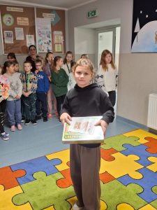 Uczennica stoi na kolorowym dywanie w dłoniach przed sobą trzyma otrzymaną w konkursie nagrodę. W tle widać stojących innych uczniów.