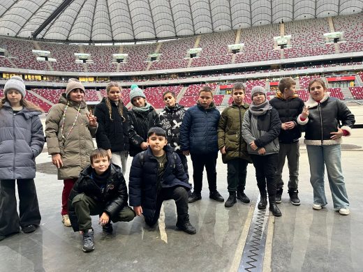 Uczniowie pozują do wspólnego zdjęcia stojąc na płycie stadionu narodowego. Dwóch chłopców kuca a reszta stoi za nimi otaczając ich w lekkim łuku. Za uczniami widoczne są trybuny stadionu.