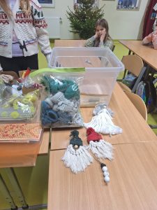 Zdjęcie ułożonych na stole trzech ozdób wykonanych z włóczki przez uczniów. Za nimi widoczne są pudełka i woreczki, z elementami z których zostały wykonane ozdoby. Przy stole dalej siedzi uczennica z głową skierowaną w stronę obiektywu.