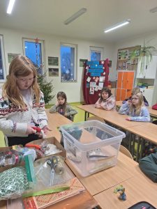Uczennica stoi przy pudełkach z przyborami do robienia ozdób świątecznych. W dłoniach trzyma jedną z ozdób, której się przygląda. Za nią przy długich stołach siedzą inni uczniowie. Przy długich połączonych ławkach za nią siedzą inni uczniowie.