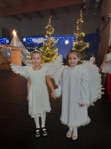 Dwie uczennice w strojach aniołków pozują do wspólnego zdjęcia na sali gimnastycznej. Dziewczynki trzymają się za ręce. W tle widoczna jest dekoracja ze światełkami.