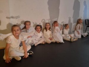 Dziewczynki z oddziału przedszkolnego przebrane za aniołki siedzą pod ścianą sali gimnastycznej obok siebie. Na ścianie widoczne są rzucane przez nie cienie.