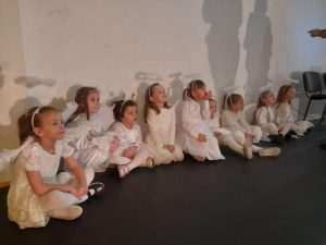 Dziewczynki z oddziału przedszkolnego przebrane za aniołki siedzą pod ścianą sali gimnastycznej obok siebie. Dziewczynki patrzą przed siebie. Na ścianie widoczne są rzucane przez nie cienie.