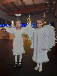 Dwie uczennice w strojach aniołków pozują do wspólnego zdjęcia na sali gimnastycznej. w tle widoczna jest dekoracja ze światełkami.