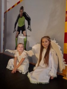 Dwie uczennice w strojach aniołków siedzą obok siebie pod ścianą sali gimnastycznej. Jedna z dziewczynek patrzy w stronę obiektywu a druga w jej kierunku. Na ścianie za nimi widoczna jest duża naklejka z bramkarzem stojącym w świetle swojej bramki.