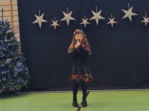 Uczennica naszej szkoły ubrana jest w czarną lekko plisowaną sukienkę oraz czarny sweterek ozdobione pasami czerwonych kwiatów. Dziewczynka w dłoniach przed sobą umieszczonych na wysokości ust trzyma mikrofon. Za dziewczynką widać ciemne tło u góry którego w jednej linii wiszą zawieszone srebrne gwiazdy w dwóch wielkościach. Obok tła widać fragment choinki.