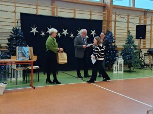 Uczennica podchodzi do kobiety trzymającej papierowe . Za nią stoi dwoje przedstawicieli jury konkursu.