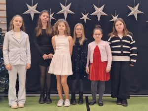 Sześć dziewczynek reprezentujących w konkursie naszą szkołę pozuje do wspólnego zdjęcia stojąc obok siebie. Za nimi na górze ciemnego tła zawieszone są srebrne gwiazdy.