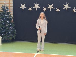 Uczennica naszej szkoły ubrana w jasny garnitur stoi wyprostowana trzymając w dłoni na wysokości pasa mikrofon. Za dziewczynką widać ciemne tło u góry którego w jednej linii wiszą zawieszone srebrne gwiazdy w dwóch wielkościach. Obok tła widać fragment choinki.