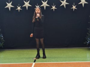 Uczennica naszej szkoły ubrana w czarną krótką sukienkę stoi trzymając w dłoni na wysokości ust mikrofon. Za dziewczynką widać ciemne tło u góry którego w jednej linii wiszą zawieszone srebrne gwiazdy w dwóch wielkościach.