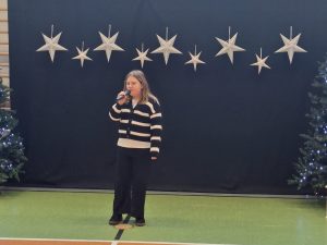 Uczennica naszej szkoły ubrana w sweter w jasno - ciemne pasy stoi na zielonej podłodze trzymając w dłoni na wysokości ust mikrofon. Za dziewczynką widać ciemne tło u góry którego w jednej linii wiszą zawieszone srebrne gwiazdy w dwóch wielkościach. Obok tła widać po obu stronach fragmenty choinek.