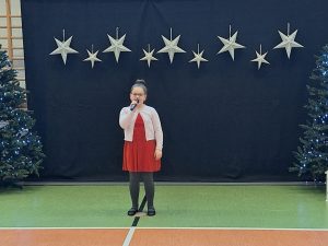 Uczennica naszej ubrana w czerwoną sukienkę i biały sweterek stoi trzymając w dłoni na wysokości ust mikrofon. Za dziewczynką widać ciemne tło u góry którego w jednej linii wiszą zawieszone srebrne gwiazdy w dwóch wielkościach. Obok tła widać po obu stronach fragmenty choinek.