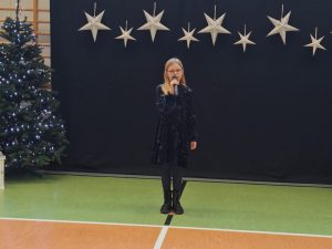 Uczennica ubrana w ciemną sukienkę stoi trzymając w dłoni na wysokości ust mikrofon. Za dziewczynką widać ciemne tło u góry którego w jednej linii wiszą zawieszone srebrne gwiazdy w dwóch wielkościach. Obok tła widać fragment choinki.