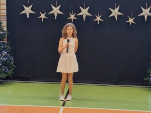 Uczennica naszej szkoły ubrana w białą sukienkę z odkrytymi ramionami stoi trzymając w jednej dłoni umieszczonej przed sobą mikrofon. Drugą rękę ma splecioną za sobą. Za dziewczynką widać ciemne tło u góry którego w jednej linii wiszą zawieszone srebrne gwiazdy w dwóch wielkościach. Obok tła widać fragment choinki.