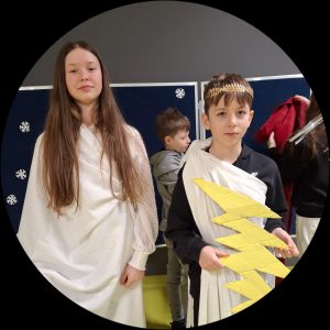 Dwoje uczniów przepasanych białymi chustami pozuje do wspólnego zdjęcia. Chłopiec na głowie ma złotą koronę a w dłoniach przed sobą trzyma wyciętą z kartonu błyskawicę.