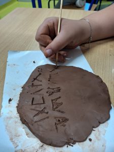 Na zdjęciu widoczna jest uklepana glina leżąca na białej podkładce oraz dłoń dziewczynki wraz z drewnianym patyczkiem przyciśniętym do gliny. Na glinie widoczne są wyżłobione znaki.