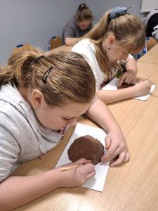 Dwie uczennice siedzą przy wspólnym stole i pochylają się nad białymi tekturkami, na których ułożone są gliniane kółka. Dziewczynki trzymają w dłoniach drewniane patyczki.