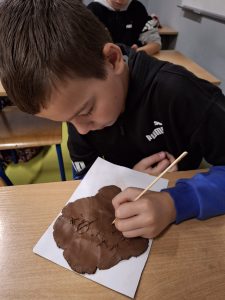 Uczeń pochyla się nad stołem, na którym leży papierowy prostokąt a na nim placek z gliny. Chłopiec rysuje na nim drewnianym patyczkiem litery.