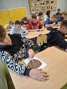 Uczniowie klasy piątej siedzą w sali przy długim stole. Przed nimi leżą podkładki z glinianymi kółkami. Chłopiec widoczny na pierwszym planie żłobi w glinie leżącej przed nim pionowe kreski.