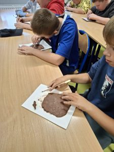 Dwóch uczniów siedzi obok siebie przy wspólnym stole. Przed nimi leżą białe podkładki z gliną. Chłopcy trzymają w dłoniach drewniane patyczki. Chłopiec widoczny na pierwszym planie uklepuje swoją glinę palcami, a jego kolega żłobi w swoim glinianym kółku patyczkiem.