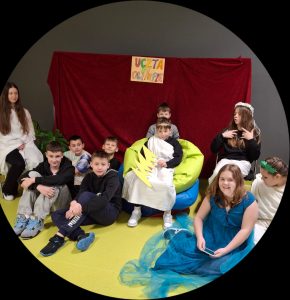 Uczniowie biorący udział w inscenizacji uczty na Olimpie pozują do wspólnego zdjęcia. Po środku na kolorowych dużych poduszkach siedzi uczeń przebrany za Zeusa trzymający w dłoniach wycięty z kartonu piorun.