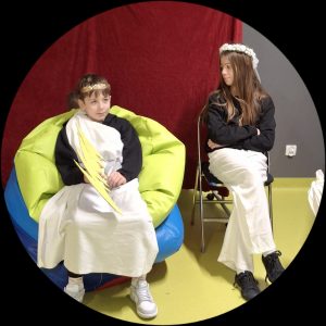 Dwoje uczniów biorących udział w przedstawieniu o greckich bogach siedzi obok siebie. Dziewczynka siedzi na krześle a na głowie ma wianek z białych kwiatów. Obok niej na wielkich kolorowych poduszkach siedzi chłopiec przebrany za Zeusa. Na głowie ma złotą koronę z liści a w dłoniach trzyma wycięty z tektury piorun.