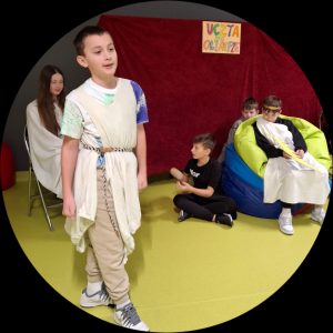 Zdjęcie scenki z przedstawienia "Uczta na Olimpie". Na pierwszym planie widoczny jest przechodzący chłopiec przepasany białym materiałem ściągniętym w pasie paskiem. Za chłopcem widoczni są inni uczniowie biorący udział w przedstawieniu.