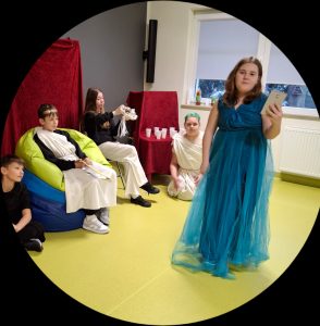 Zdjęcie sceny z przedstawienia pt. "Uczta na Olimpie". Na pierwszym planie widoczna jest uczennica ubrana w długą niebieską suknię i trzymająca w dłoni na wysokości oczu mały notesik. Za nią siedzą inni uczniowie biorący udział w przedstawieniu. W centralnym miejscu siedzi na kolorowych pufach chłopiec przebrany za Zeusa. Dziewczynka siedząca na krześle obok trzyma w dłoniach biały wianek, a dalej na podłodze siedzi uczennica ubrana na biało z zielonym wiankiem na głowie.