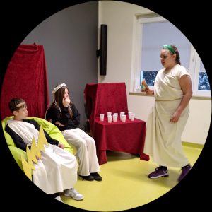 Zdjęcie sceny z "Uczty na Olimpie". Do dwojga uczniów siedzących na sali podchodzi ich koleżanka przepasana białym materiałem udrapowanym na pasku w pasie. Dziewczynka w dłoni trzyma jakiś przedmiot. Obok niej na stole zakrytym czerwonym materiałem stoją białe plastikowe kubeczki. dwoje uczniów siedzących naprzeciwko niej ubranych jest w stroje bogów.