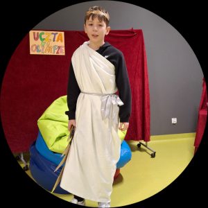 Uczeń samotnie stoi na sali. Za nim widoczna jest tablica zakryta czerwonym materiałem i z przyczepioną u góry kartką z napisem "UCZTA NA OLIMPIE". Chłopiec przepasany jest białym materiałem związanym w pasie biała wstążką. Na głowie ma złotą koronę z liści. A w dłoni u swojego boku trzyma wycięty z tektury piorun.