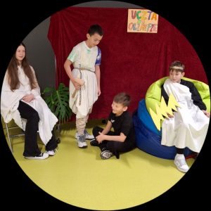 Uczniowie klasy piątej podczas inscenizowanego przedstawienia. Na zdjęciu widać czterech uczniów. Patrząc od lewej strony na krześle siedzi uczennica przepasana białym materiałem. Obok niej stoi chłopiec również przepasany materiałem z zapiętym na nim paskiem i trzymający w dłoni drewniany młotek. Chłopiec pochyla się nad siedzącym na podłodze kolegą ubranym na czarno. Po prawej stronie na kolorowych dużych pufach siedzi uczeń przebrany za Zeusa trzymający w dłoniach wycięty z tektury piorun.