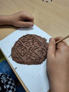 Na zdjęciu widoczna jest uklepana glina leżąca na białej podkładce oraz dwie dłonie chłopca wraz z drewnianym patyczkiem trzymanym przez jedną z nich i przyciśniętym do gliny. Na glinie widoczne są wyżłobione znaki.