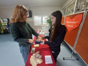 Nauczycielka wybiera z pudełka los biorący udział w loterii. Na przeciwko niej stoi uczennica trzymająca pudełko. Pomiędzy nimi ustawiony jest czerwony stół na którym leżą przygotowane na loterię fanty.