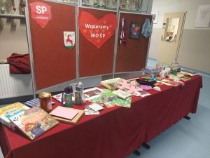 Zdjęcie stołu przykrytego czerwonym materiałem, na którym zostały ustawione przedmioty przygotowane na loterię fantową. Za stołem na czerwonej tablicy zostały umieszczone logo szkoły i gminy oraz duże serce z napisem "Wspieramy WOŚP".