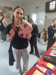 Uczennica klasy siódmej pozuje do zdjęcia trzymając na wysokości twarzy los oraz wygraną nagrodę - różową podkładkę w kształcie kwiatka. W tle widać innych uczniów chodzących po szkolnym korytarzu.