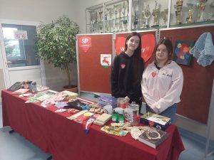 Dwie uczennice sprzedające losy w loterii pozują do wspólnego zdjęcia stojąc za długim stołem na którym ułożone zostały fanty na loterię.