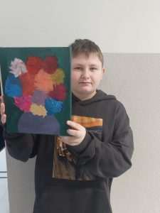 Uczeń klasy szóstej pozuje do zdjęcia trzymając na wysokości twarzy wygrany w loterii obraz.