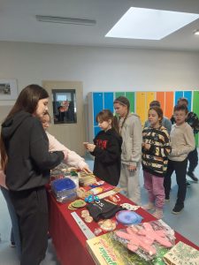 Uczniowie klasy czwartej stoją w rzędzie przed stołem, na którym leżą rozłożone fanty przygotowane na loterię. Na przeciwko nich stoją dwie uczennice sprzedające losy. Jedna z nich wskazuje placem na przedmiot leżący na stole.