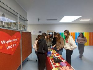 Uczennice klasy siódmej stoją przy stoisku, gdzie są rozłożone fanty przygotowane na loterię. Dziewczynki im się przyglądają. Po drugiej stronie stołu stoją uczennice sprzedające locy.