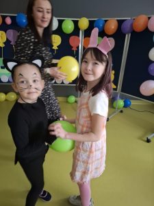 Dwie uczennice stoją na przeciwko siebie trzymając się za dłonie. Pomiędzy ich brzuchami jest umieszczony balon, który podtrzymują swoimi ciałami. W tle widoczna jest pani nauczycielka trzymająca w dłoniach balon i spoglądająca w stronę dziewczynek.