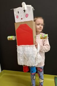 Uczennica oddziału przedszkolnego pozuje do zdjęcia trzymając przed sobą wykonanego na konkurs robota. Figurka zasłania połowę jej ciała i wystaje ponad jej głowę. Zdjęcie wykonane na tle czarnej ściany.