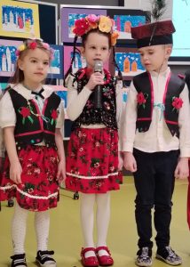 Troje uczniów oddziału przedszkolnego w strojach krakowiaków stoi blisko obok siebie. Dziewczynka stojąca po środku ma trzyma na wysokości ust mikrofon.