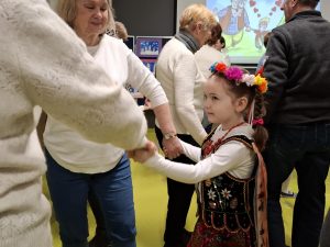 Uczennica oddziału przedszkolnego ubrana w strój krakowianki tańczy z dwoma babciami, które trzyma za dłonie. W tle widać inne tańczace osoby.