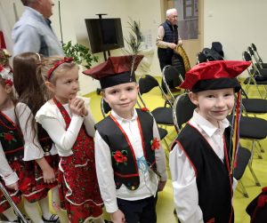 Uczniowie oddziału przedszkolnego ubrani w stroje krakowiaków stoją w rzędzie. Za nimi stoją ustawione w rzędy puste krzesła. Dwóch chłopców widocznych na pierwszym planie uśmiecha się w stronę obiektywu.