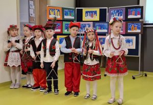 Siedmioro uczniów oddziału przedszkolnego stoi w jednym rzędzie. Dzieci ubrane są w stroje krakowiaków a w dłoniach przed sobą trzymają trójkąty do grania.