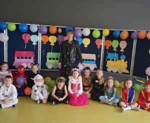 Uczniowie oddziału przedszkolnego siedzą obok siebie, w swoich kostiumach na podłodze sali teatralnej. za nimi stoi ich wychowawczyni. Za nią widoczna jest tablica ozdobiona kolorowymi, nadmuchanymi balonami oraz wyciętym z kolorowego papieru pociągiem.