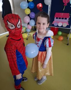 Dwoje uczniów stoi na przeciwko siebie z balonem , który podtrzymują swoimi brzuchami. Dziewczynka ubrana jest w strój księżniczki a chłopiec przebrany jest za Spider-Mena.