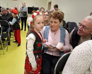 Uczennica oddziału przedszkolnego stoi w stroju krakowianki obok siedzących na krzesełkach dziadków. Babcia trzyma w dłoni otrzymane od dziewczynki podarki, a dziadek pochyla się w stronę swojej wnuczki. Dziewczynka patrzy w stronę obiektywu.