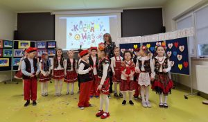 Uczniowie oddziału przedszkolnego w strojach krakowiaków pozują do wspólnego zdjęcia. Z tyłu stoją dziewczynki w jednym rzędzie wraz z umiejscowioną po środku wychowawczynią. Z przodu w nieregularny sposób ustawieni są chłopcy. Przed nimi w centralnym punkcie stoi bokiem ich koleżanka. W tle na rzutniku wyświetlony jest kolorowy napis "kochamy was".
