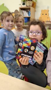 Uczennica oddziału przedszkolnego pozuje do zdjęcia trzymając w dłoniach przed sobą otrzymane nagrody - pudełko z pisakami i drugie z matematyczną łamigłówką. Opakowania przysłaniają dziewczynce dolną część jej twarzy. Za nią widoczne są dwie jej koleżanki stojące blisko siebie i patrzące w stronę obiektywu.